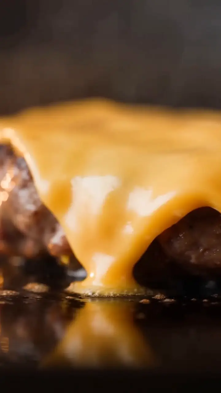 Still from a burger commercial: melted cheese sliding over a seared patty on the grill, close up.